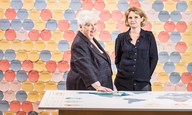 Barbara and Sophie, both dressed in black, stand between a table with a map of the world and a wall of coloured (blue, yellow, red and pink) discs on which exhibition visitors have written their own or their family's migration or non-migration stories.